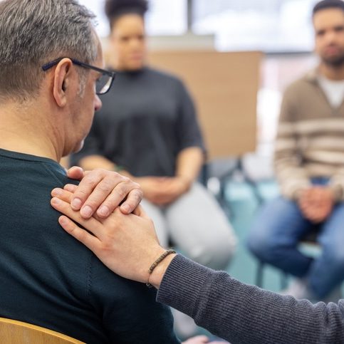 Participant places a hand on the shoulder of an upset senior man during a group therapy session, offering emotional support in a caring community setting. Senior man gets emotional support from another participant during a group therapy session at a community center.